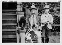 Man holding gun, seated next to young boy and two dogs.