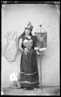 Woman with heart-shaped breastplate and spectacle lenses hanging on costume holds a sign for F. T. Bessac.