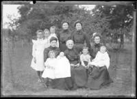 Group of 5 women, 3 girls, 3 boys, and baby standing and sitting in yard